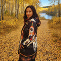 A woman wearing a black hooded jacket with a colorful graphic print stands on a leaf-covered path in an autumn forest with birch trees and a river in the background.